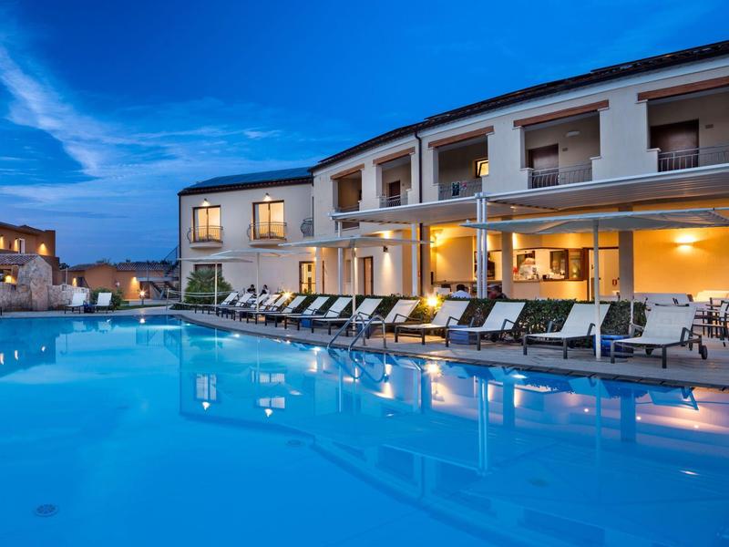 Hotel mit beleuchtetem Außenpool und Liegestühlen am Abend unter blauem Himmel.