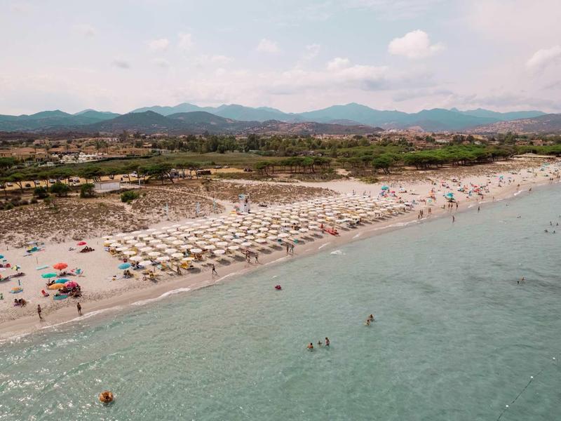 Lange zandstrand met ligbedden en parasols, duinen en bergen op de achtergrond onder een bewolkte lucht.