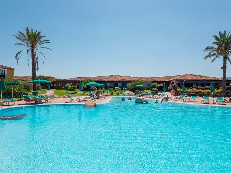 Groot hotelzwembad met ligstoelen en palmbomen onder een zonnige hemel.