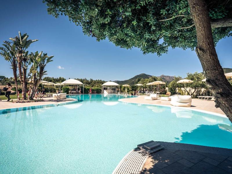 Large hotel pool with umbrellas and palm trees under clear sky.