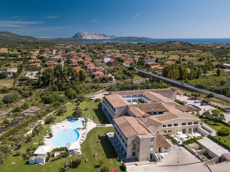 Aerial view of a hotel with swimming pool, surrounded by greenery and mountains in the background.