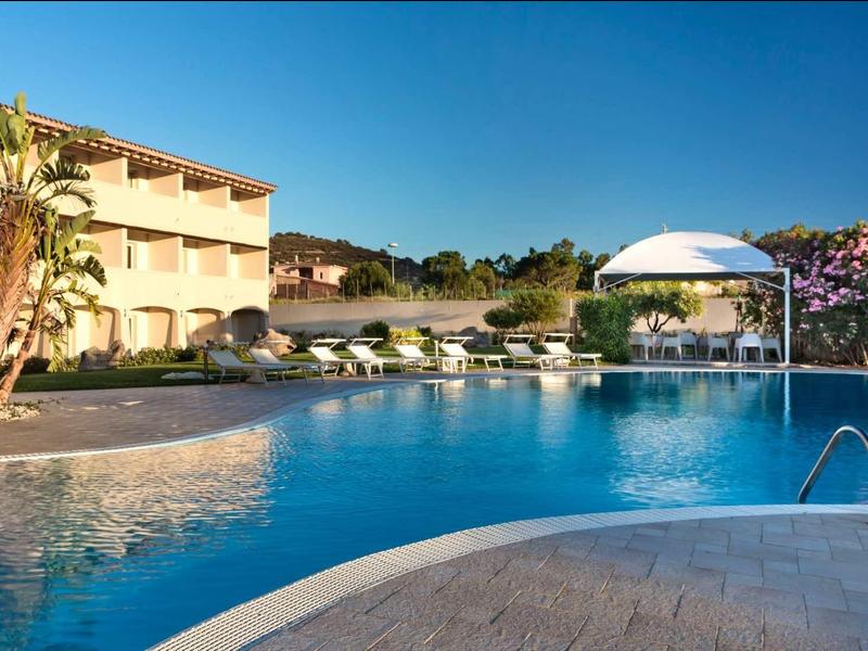 Large hotel pool with sun loungers and shade, surrounded by palm trees and flowering bushes.