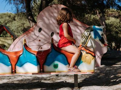 Kind in roter Kleidung sitzt auf buntem Schaukeltier aus Holz, im sonnigen Park mit Bäumen.
