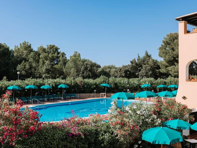 Ein klarer Pool umgeben von grünen Büschen, blühenden Blumen und blauen Sonnenschirmen bei klarem Himmel.