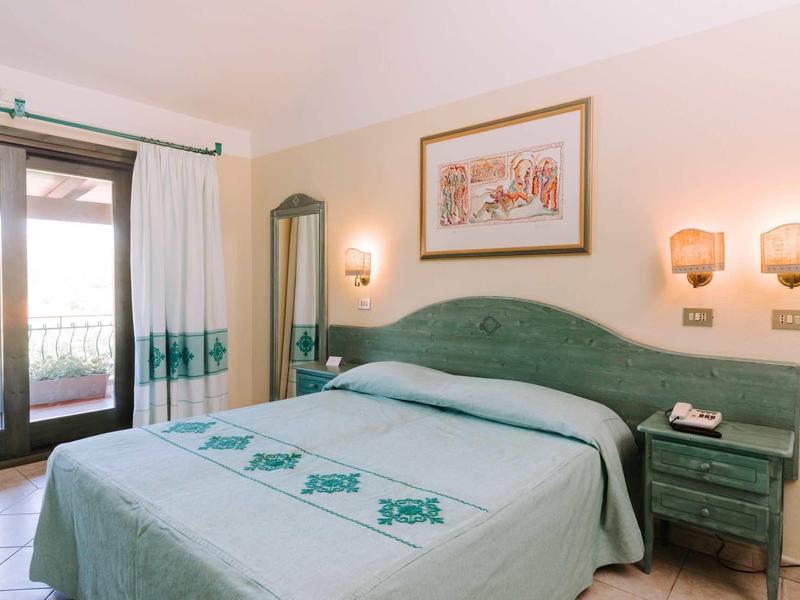 Hell eingerichtetes Schlafzimmer mit grünem Bett, Nachttisch, Bilderrahmen und Balkonzugang.