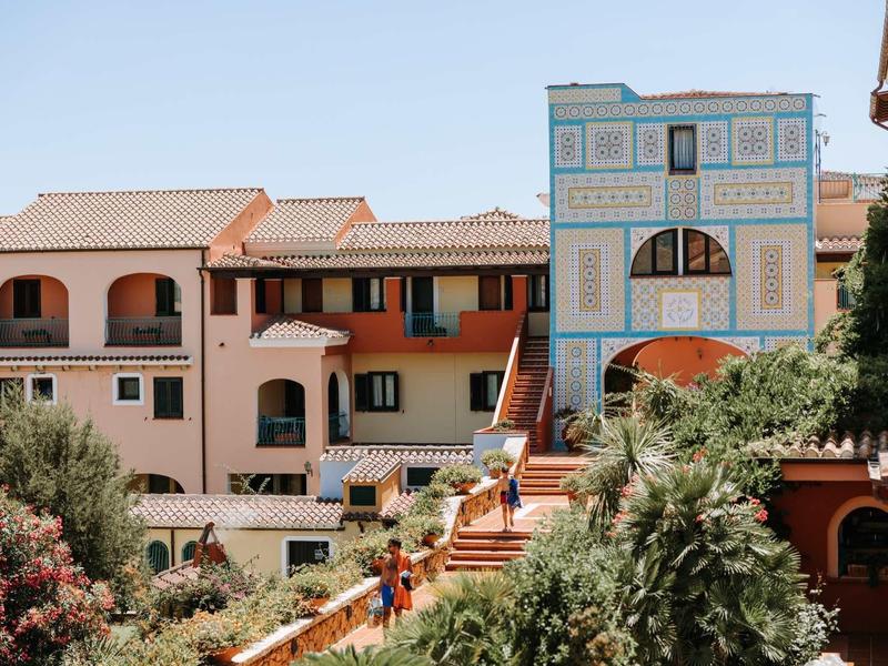 Bunte mediterrane Häuser mit Bögen und Balkonen, umgeben von grünen Pflanzen unter klarem Himmel.