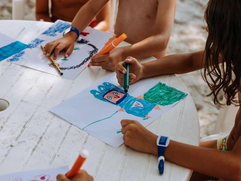 Kinder zeichnen mit bunten Filzstiften auf weißem Tisch, Sommergefühl, helle Holzstruktur sichtbar.