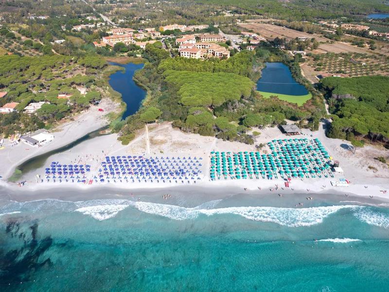 Luftaufnahme eines Strandes mit blauen und grünen Sonnenschirmen, türkisfarbenem Meer und grüner Landschaft.