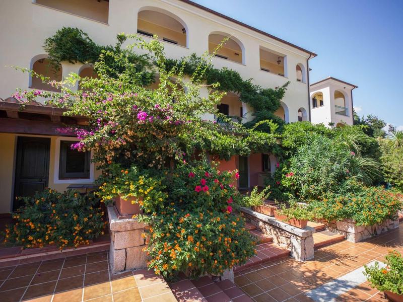 Großes Haus mit weißem Putz, Pflanzen und Bougainvillea, Terrasse mit Rattanmöbeln und roten Fliesen.