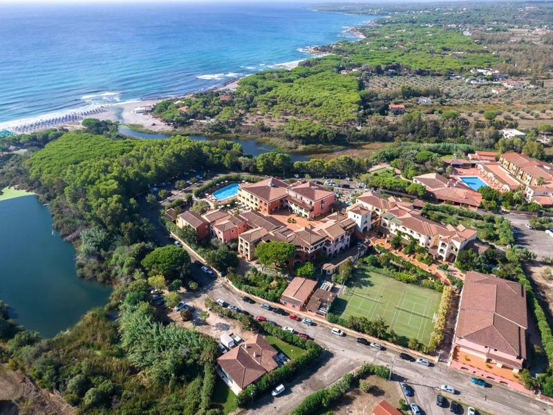 Luftaufnahme einer Küstenregion mit Gebäuden, Grünflächen, einem See und blauem Meer im Hintergrund.
