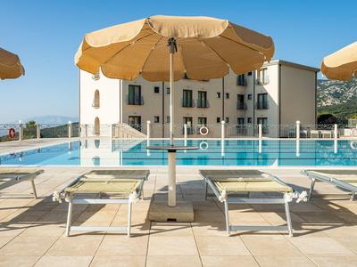 Piscina con lettini e ombrelloni davanti a un edificio dell'hotel sotto un cielo limpido.