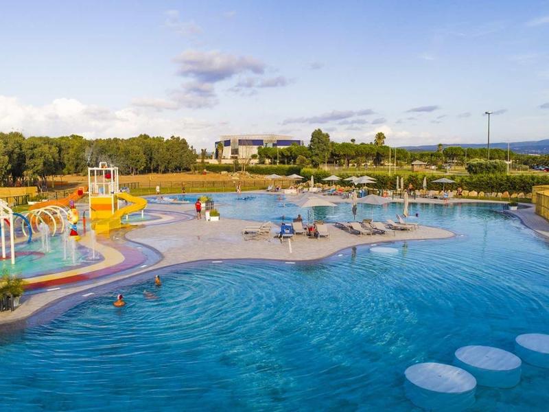 Grand bassin extérieur avec jeux d'eau et espaces de repos, entouré de verdure et ciel bleu.