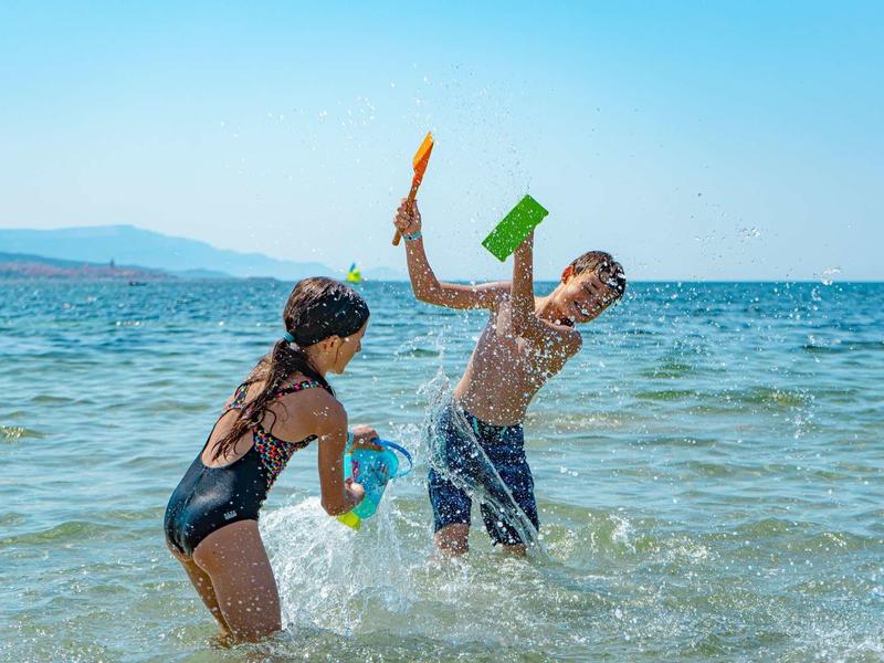 Deux enfants jouent avec des seaux et des pelles dans l'eau peu profonde de la mer sous un ciel clair.