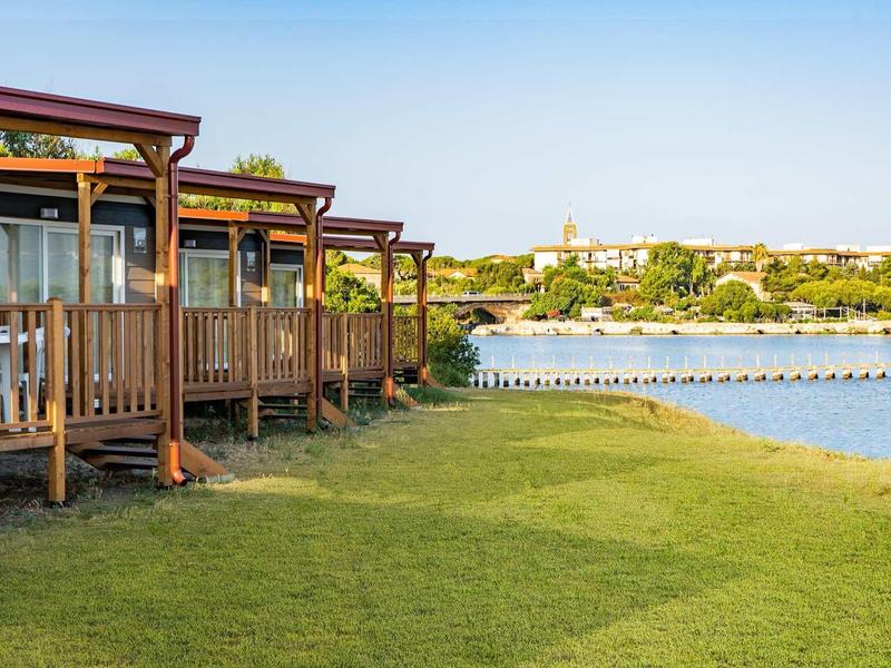Chalets en bois au bord d'un lac calme avec de l'herbe verte et un ciel clair.