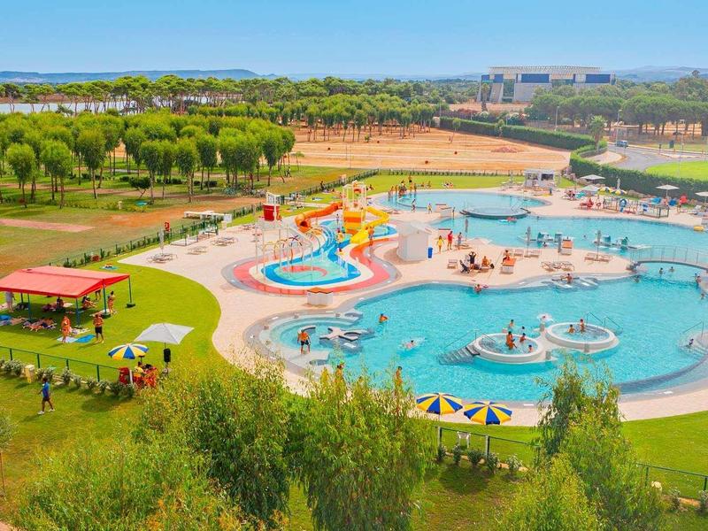 Une grande piscine avec des toboggans aquatiques et des personnes, entourée de pelouses vertes et d'arbres par temps ensoleillé.