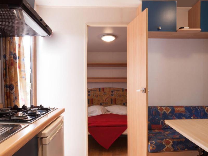 Kompakte Wohnküche mit Gasherd und Sitzbereich neben einem Schlafzimmer mit roten Bettdecken.