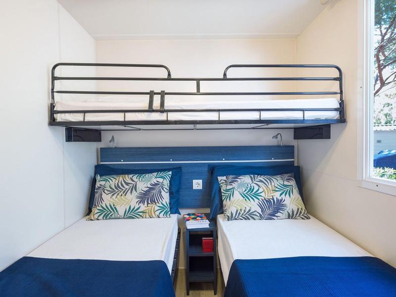 Kleines Schlafzimmer mit Etagenbett, zwei Einzelbetten und einem Fenster mit Blick ins Grüne.