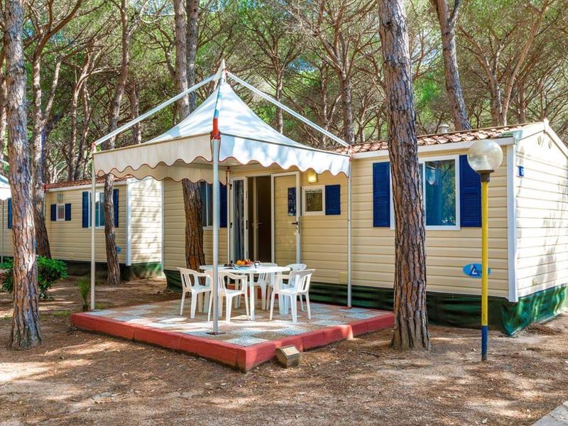 Mobilheim mit weißem Sonnensegel und Außenmöbeln in einem bewaldeten Ferienpark.
