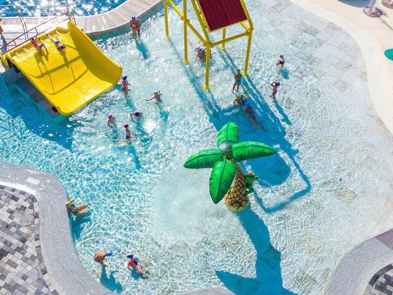 Wasserpark mit Pool, Rutschen und schattenspendenden Sonnenschirmen, belebt mit spielenden Menschen.