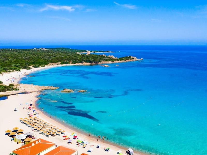 Strand mit klarem türkisfarbenem Wasser, Sonnenschirmen und grüner Küstenlandschaft.