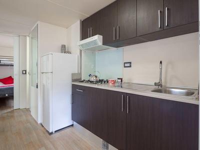 Malá moderní kuchyň s tmavými skříňkami, sporákem, lednicí a výhledem do ložnice.