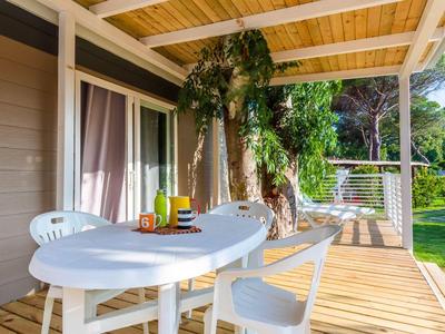 Dřevěná terasa s bílým stolem a židlemi, ovocem a nápoji, obklopená zelení.