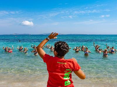 Persoon in rood shirt staat op het strand en zwaait naar een groep mensen in het water.
