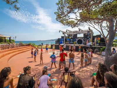 Mensen dansen voor een openluchttheater met uitzicht op zee en omringd door bomen.