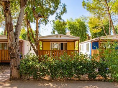 Drei kleine Holzbungalows mit Balkonen, umgeben von grünen Büschen und Bäumen bei Sonnenschein.