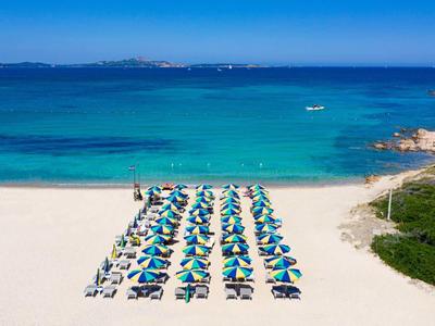 Pláž s řadami lehátek a slunečníků u čistého modrého moře a zalesněného pobřeží.