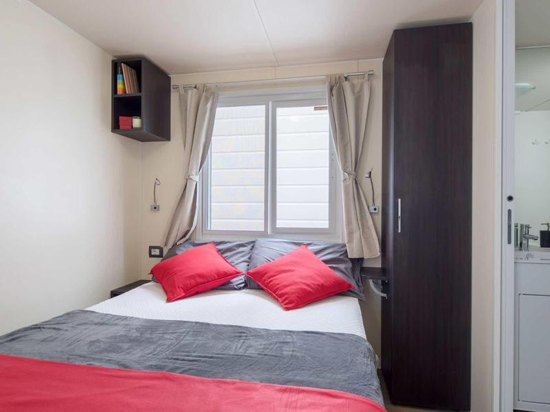 Malá ložnice s manželskou postelí, šedým povlečením, červenými polštáři a malou skříní.