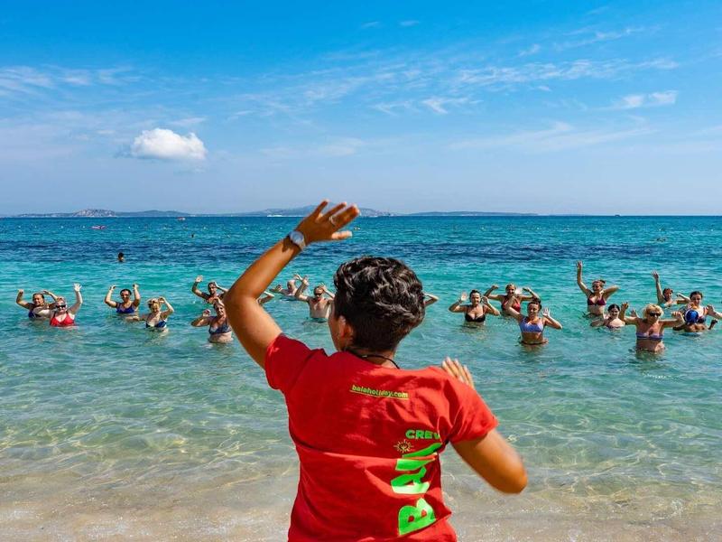 Žena v červeném tričku vede vodní aerobik v průzračném moři s účastníky.