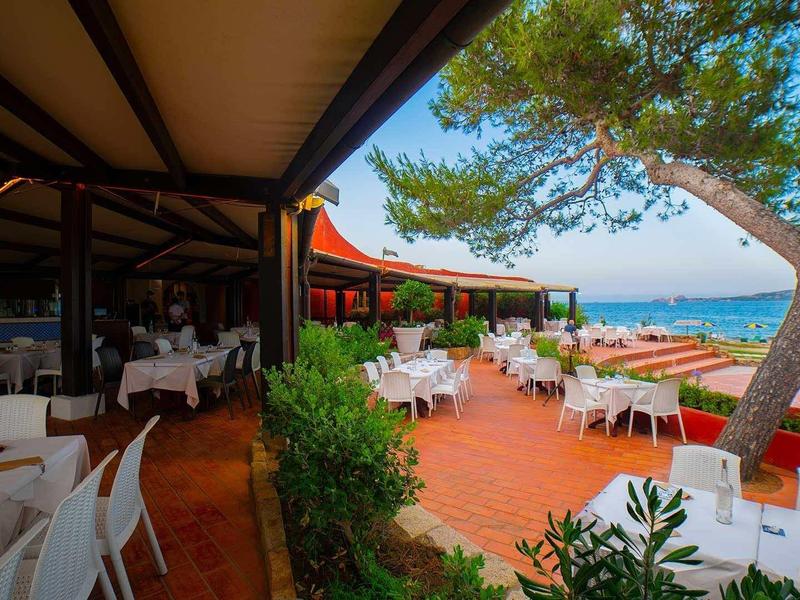 Terasa restaurace s bílými stoly a židlemi s výhledem na moře a stromy.