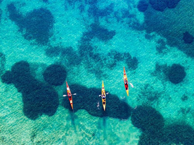 Tři kajakáři pádlí v průzračné tyrkysové vodě s tmavými stíny pod sebou.