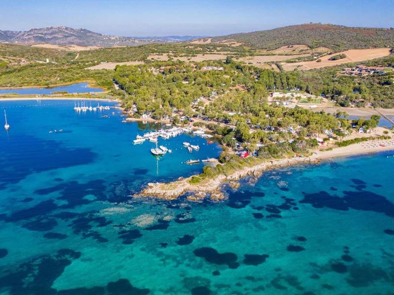 Vista aerea della costa con acqua blu chiara, barche a vela e riva boscosa.