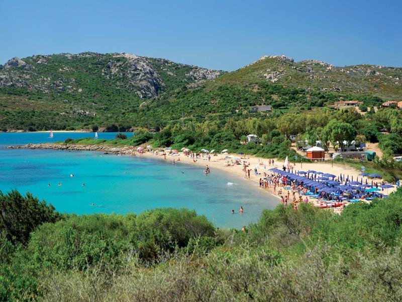 Spiaggia con acqua azzurra limpida, ombrelloni e montagne sullo sfondo.