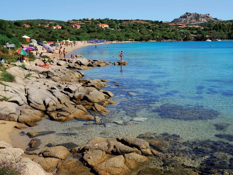 Paesaggio costiero con acqua blu chiara, rocce e spiaggia sabbiosa in una giornata soleggiata.