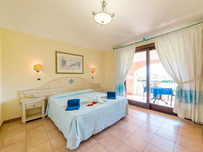 Helles Schlafzimmer mit Doppelbett, zwei Nachttischen, Lampen, Vorhängen und Balkon mit blauem Geländer.