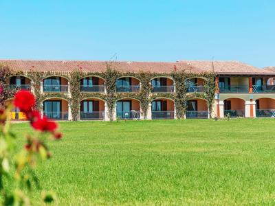 Ein zweistöckiges Gebäude mit Rundbogenfenstern, davor ein großer grüner Rasen und blühende rote Blumen.