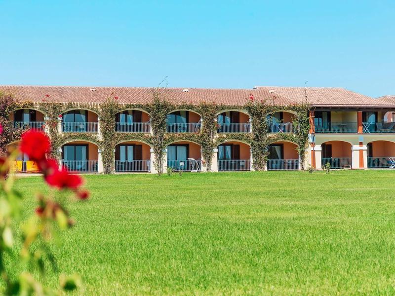 Ein zweistöckiges Gebäude mit Rundbogenfenstern, davor ein großer grüner Rasen und blühende rote Blumen.