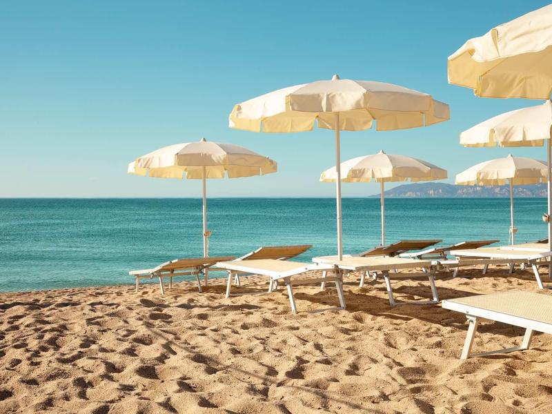 Strand mit Sand, mehreren weißen Sonnenschirmen und Liegestühlen unter klarem Himmel.
