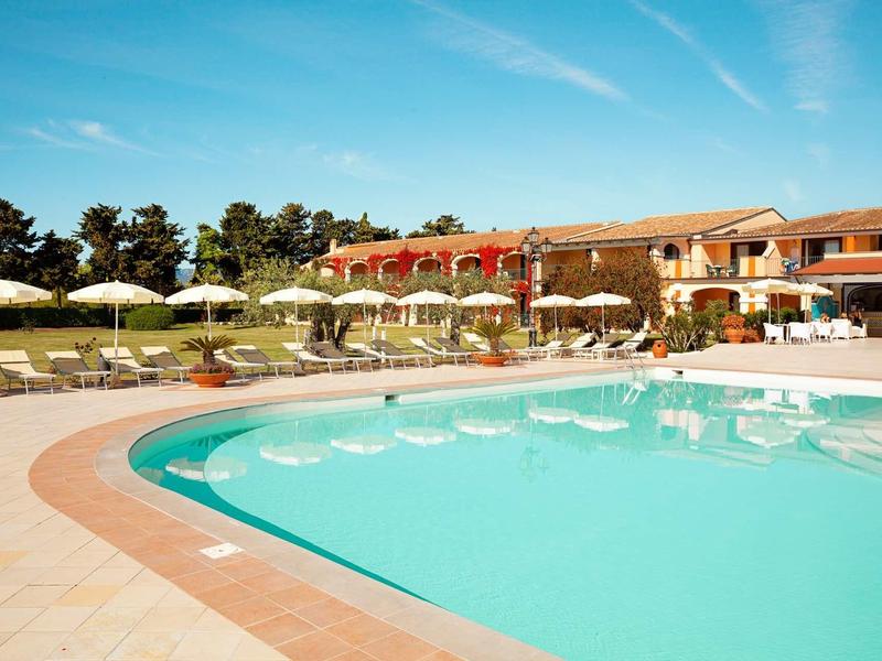 Großer Pool mit türkisblauem Wasser, Sonnenschirmen und roten Gebäuden im Hintergrund bei klarem Himmel.