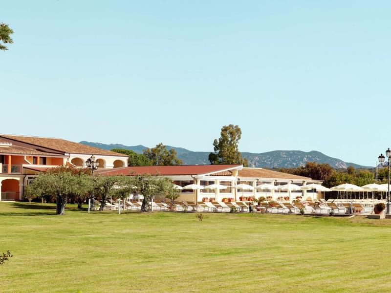 Großes Hotel mit rotem Dach vor grüner Wiese und Bergen im Hintergrund unter klarem Himmel.