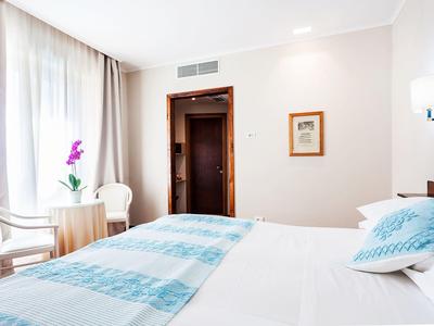 Jasny pokój hotelowy z podwójnym łóżkiem, dwoma krzesłami, dużym oknem i rośliną.