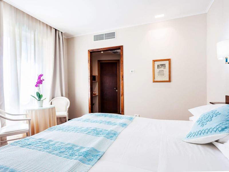 Jasny pokój hotelowy z podwójnym łóżkiem, dwoma krzesłami, dużym oknem i rośliną.