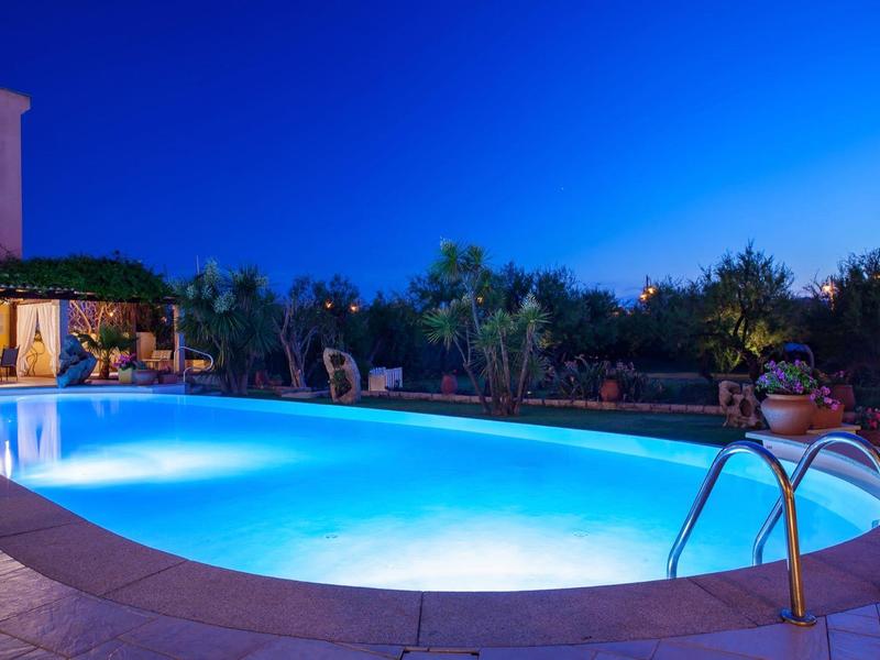 Oświetlony basen nocą z schodkami otoczony roślinami i miejscami do siedzenia.