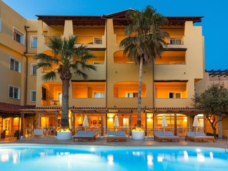 Budynek hotelu o zmierzchu z oświetlonym basenem i palmami z przodu.