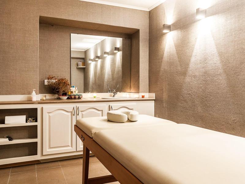 Jasny pokój do masażu z białym stołem, ręcznikami i dużym lustrem na ścianie.