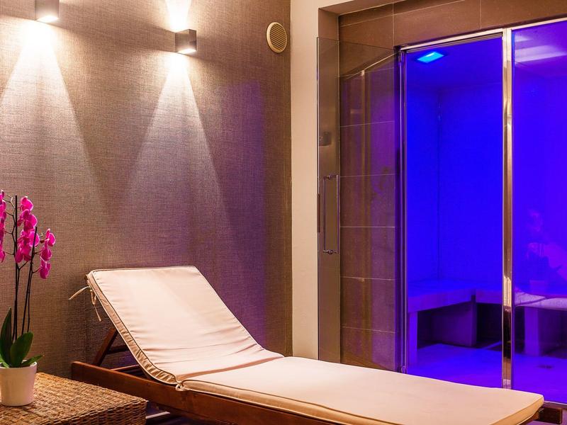 Pokój relaksacyjny z wygodnym leżakiem, kwiatem i niebiesko podświetlonym szklanym drzwiom do strefy wellness.