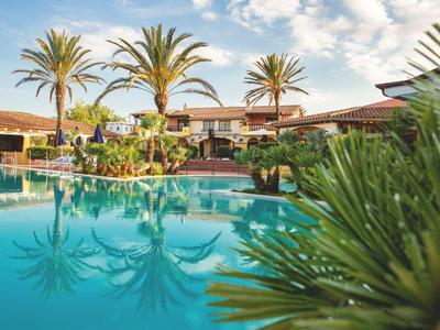 Velký bazén s palmami a hotelem v pozadí pod částečně oblačnou oblohou.